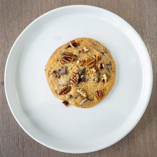 A chocolate chip pecan cookie on a white plate, set against a wooden table.