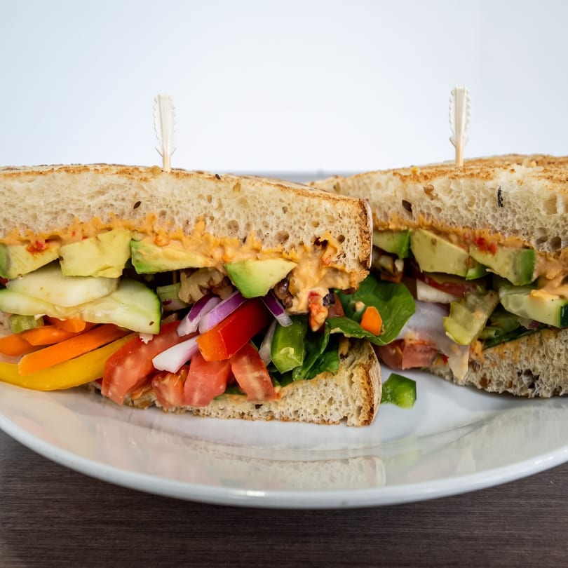 Two halves of a hearty sandwich filled with colorful vegetables, avocado, and a creamy sauce.